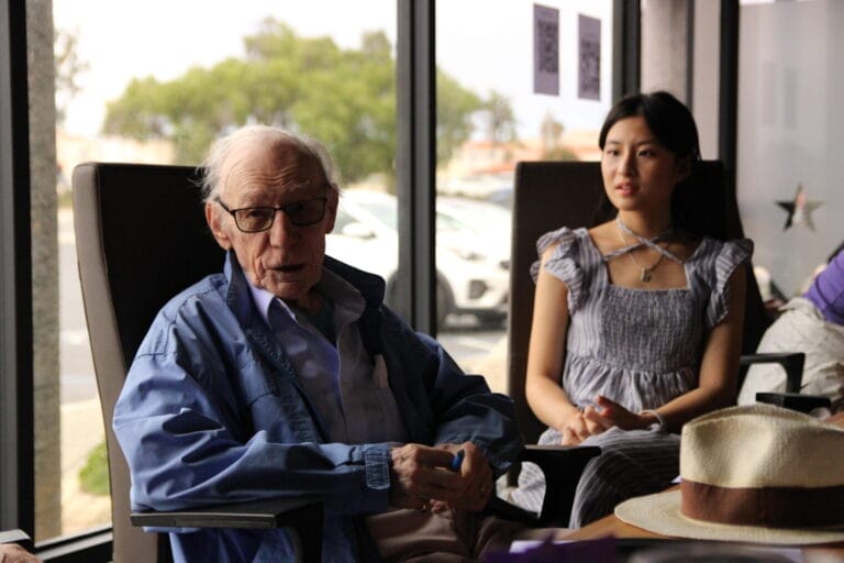 About | curaJOY An elderly man in glasses and a blue jacket sits next to a young woman in a gray dress inside a room with large windows, reflecting a warm moment that highlights the importance of emotional wellness and connection across generations.