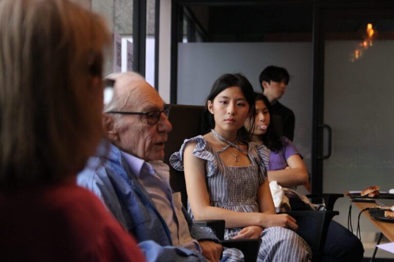 About | curaJOY A group of people sit indoors, engaged in conversation about emotional wellness; one older man speaks while a young woman in a striped dress listens attentively, with others in the background, reflecting the spirit of curajoy.