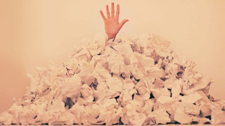 A single hand reaches upward from a large pile of crumpled white paper sheets, suggesting healthcare providers overwhelmed or buried in work.