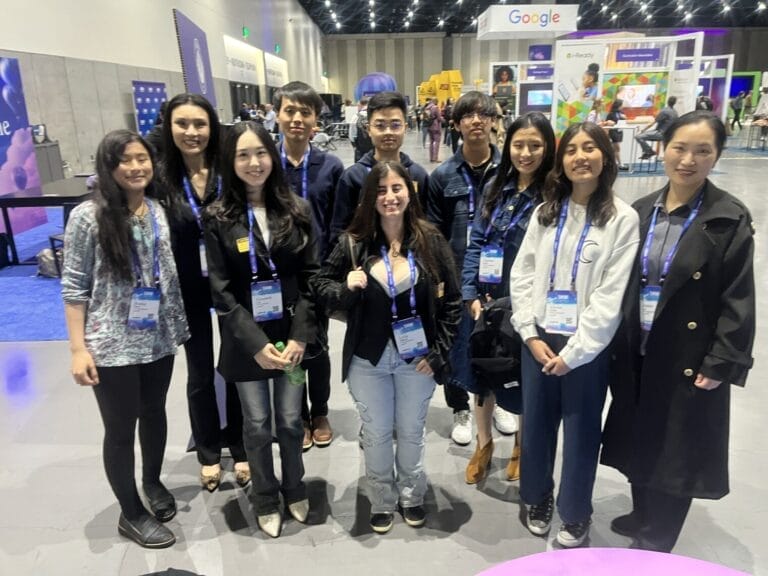 Homepage | curaJOY A group of ten people wearing conference badges pose together indoors at a tech event, with booths and displays visible in the background.